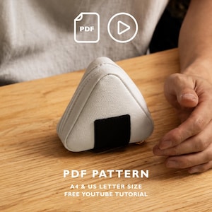 Op de afbeelding: Een driehoekig wit tasje met textuur en een zwart rechthoekig accent, dat lijkt op een onigiri. De afbeelding bevat iconen voor een PDF en een video, met de tekst PDF PATTERN, A4 & US LETTER SIZE en FREE YOUTUBE TUTORIAL.