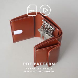 May include: A brown leather key holder wallet with white stitching, featuring a metal six-hook key rack inside. The image includes icons for PDF and video, with text reading PDF PATTERN, A4 & US LETTER SIZE, FREE YOUTUBE TUTORIAL.