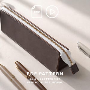 Peut inclure: Une trousse rectangulaire marron foncé avec une fermeture éclair blanche et des dents en métal doré. Le texte indique PDF PATTERN, A4 & US LETTER SIZE, FREE YOUTUBE TUTORIAL. Deux stylos sont visibles à proximité, un clair et un foncé.