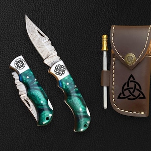 May include: A set of three items: a folding knife, a smaller folding knife, and a brown leather sheath. The knives have green and white handles with a Celtic knot design. The knife blades have a patterned design.