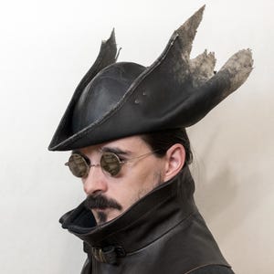 May include: A black leather tricorn hat with a distressed edge. The hat has a high crown and a wide, upturned brim. The hat is worn with round, wire-rimmed glasses and a black leather vest. The hat's brim is torn.