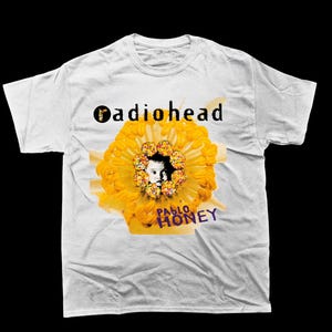 May include: White t-shirt featuring the band name "Radiohead" in black text above a yellow flower design with a face in the center. The words "Pablo Honey" are printed in purple below the flower.