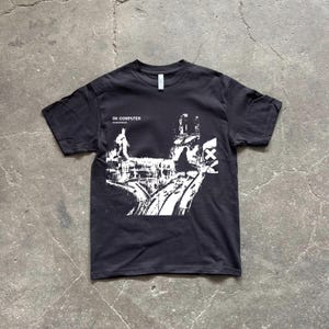 Può includere: T-shirt nero con un design grafico bianco e la scritta "OK COMPUTER". Il design raffigura un paesaggio urbano stilizzato. La maglietta ha un girocollo ed è in tessuto morbido.