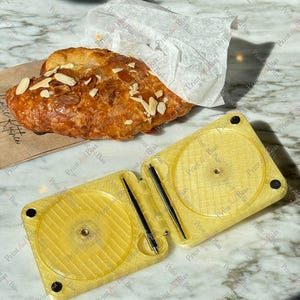 May include: A golden yellow, rectangular, hinged plastic case with two circular recessed areas and two thin black metal rods inside. Next to the case is a flaky, golden-brown almond croissant topped with sliced almonds, partially wrapped in white paper.