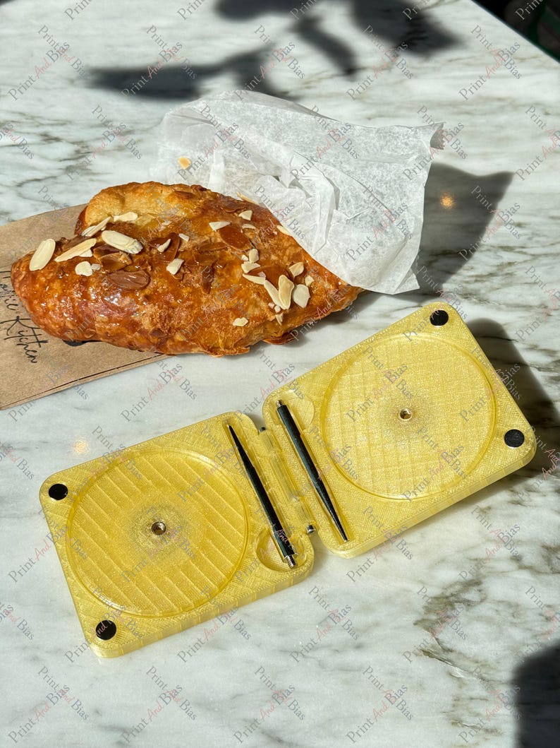 May include: A golden yellow, rectangular, hinged plastic case with two circular recessed areas and two thin black metal rods inside. Next to the case is a flaky, golden-brown almond croissant topped with sliced almonds, partially wrapped in white paper.