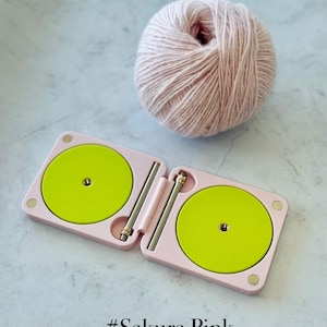 May include: A pink double compact with two bright green circles and silver metal rods. A ball of light pink yarn sits next to the compact. The text "#Sakura Pink Double Compact" is at the bottom.
