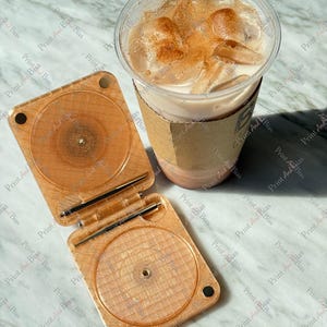 May include: A light brown, square, hinged plastic case with a glittery finish and circular indentations, paired with a clear plastic cup containing an iced beverage topped with a dusting of brown powder and a tan cardboard sleeve.