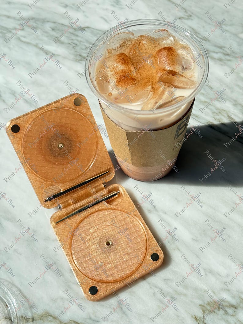 May include: A light brown, square, hinged plastic case with a glittery finish and circular indentations, paired with a clear plastic cup containing an iced beverage topped with a dusting of brown powder and a tan cardboard sleeve.