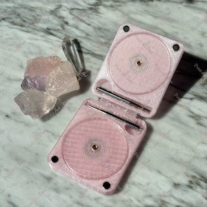 May include: A light pink, hinged, square-shaped plastic case with two circular recessed areas and metal hinge pins, positioned next to a small collection of raw, translucent, light pink and purple crystal stones on a white marble surface.