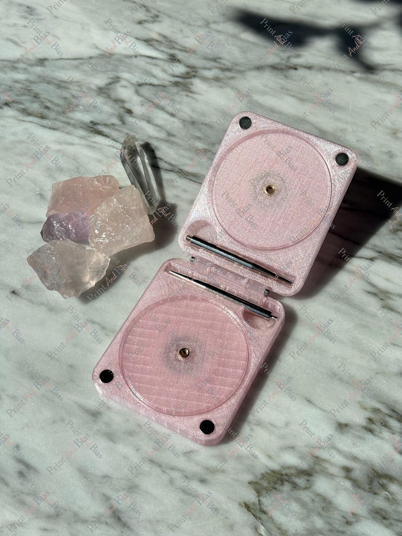 May include: A light pink, hinged, square-shaped plastic case with two circular recessed areas and metal hinge pins, positioned next to a small collection of raw, translucent, light pink and purple crystal stones on a white marble surface.