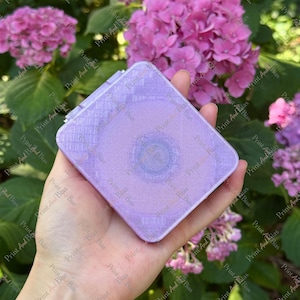 May include: A square, lavender-coloured compact mirror with a subtle, grid-like textured pattern on its surface. The compact features a smooth, slightly iridescent finish and a circular indentation in the centre, reflecting a soft, light purple hue.