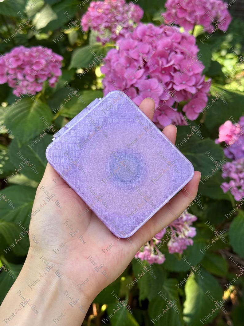 May include: A square, lavender-coloured compact mirror with a subtle, grid-like textured pattern on its surface. The compact features a smooth, slightly iridescent finish and a circular indentation in the centre, reflecting a soft, light purple hue.