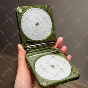 May include: A square, olive green, textured folding case with rounded corners, featuring two circular, silver-toned, mesh-like inserts with central brass-coloured threaded holes. A thin, metallic rod is stored horizontally in the centre hinge.