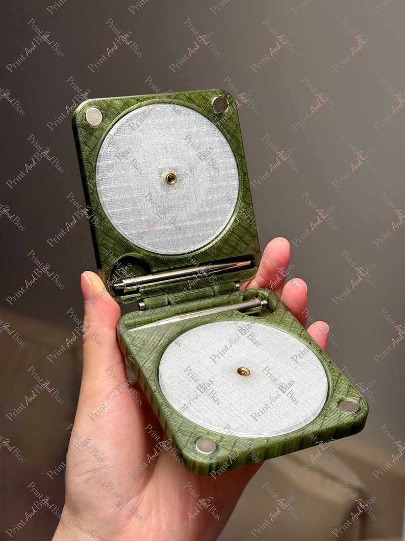 May include: A square, olive green, textured folding case with rounded corners, featuring two circular, silver-toned, mesh-like inserts with central brass-coloured threaded holes. A thin, metallic rod is stored horizontally in the centre hinge.