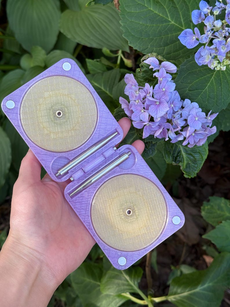 May include: A lavender, square-shaped folding case with a textured surface, featuring two circular, translucent gold-toned inserts. The case is connected by a central hinge with two silver metal rods and includes small circular magnets at the corners.