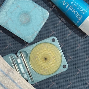 May include: A light blue, square, 3D-printed case with a textured surface, shown both closed and open. The open case reveals a circular yellow insert and two silver metal tools. Beside it is a tube of Biore UV Aqua Rich Watery Gel sunscreen.