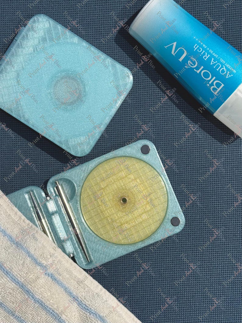 May include: A light blue, square, 3D-printed case with a textured surface, shown both closed and open. The open case reveals a circular yellow insert and two silver metal tools. Beside it is a tube of Biore UV Aqua Rich Watery Gel sunscreen.