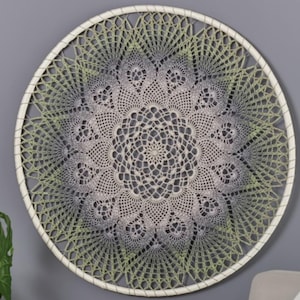 May include: A circular dreamcatcher with a cream-coloured frame and intricate crocheted web. The web has a gradient effect, transitioning from grey to green. The central design is a detailed, lacy pattern, creating a decorative wall hanging.