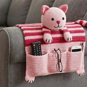 May include: A pink and red striped crochet couch organizer with a cat design. The organizer has three pockets for a remote, glasses, and a phone. The cat has a pink head, paws, and tail.