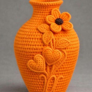 Op de afbeelding: Oranje gehaakte vaas met een bloemen- en hartontwerp. De handgemaakte vaas heeft een getextureerd oppervlak en een decoratief bloemen- en hartmotief, wat een vleugje handgemaakte charme toevoegt. De vaas is ongeveer 20 cm hoog.