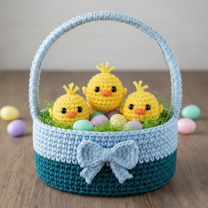 Può includere: Un cestino pasquale all'uncinetto nei toni del blu e del verde acqua, ornato da un fiocco. All'interno, tre pulcini gialli all'uncinetto sono posizionati su erba finta verde, circondati da uova di Pasqua dai colori pastello. Il cestino ha un manico.