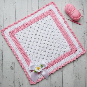 May include: A handmade, square crochet blanket in white, pink, and white. The blanket features a scalloped edge, a pink border, and a white center with a floral bow. A ball of pink yarn and a crochet hook are in the background.