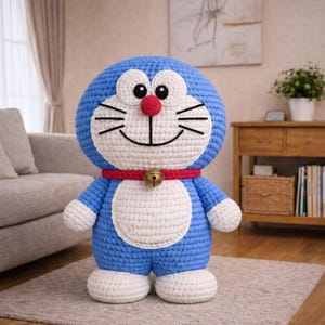 May include: A handmade blue and white crochet figure of a cartoon cat with a red collar and bell. The figure has a large head, white belly, and white feet. The figure is standing on a gray rug.