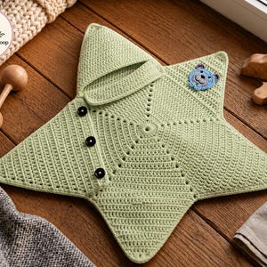 May include: A light green star-shaped crochet blanket featuring a hood, three black buttons along one side, and a small blue bear face appliqué. The item is textured with a consistent stitch pattern and includes a circular logo with the text CraftDesignsLoop.
