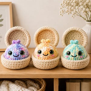 Puede incluir: Tres criaturas de peluche tejidas a ganchillo, redondas, con grandes ojos negros y pequeñas sonrisas, cada una dentro de un recipiente beige, abierto y circular, también tejido a ganchillo. Las criaturas presentan hilos jaspeados en tonos violeta, naranja y turquesa.
