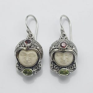 May include: A pair of silver earrings featuring a carved face design. Each earring has a small red gemstone at the top, a carved face in the center, and a green gemstone at the bottom. The earrings are on a white background.