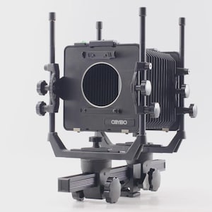 以下が含まれることがあります： 黒色の大型カメラ。蛇腹、ノブ、レンズ開口部があります。カメラは、調整可能なレールとサポートを備えたベースに取り付けられています。「OSYBO」のブランド名が前面に表示されています。