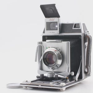 Puede incluir: Una cámara plateada y negra vintage con una lente grande y un fuelle. La cámara tiene un visor montado en la parte superior y un asa de cuero. La lente está marcada "LINHOF". La cámara está abierta, revelando sus componentes internos.