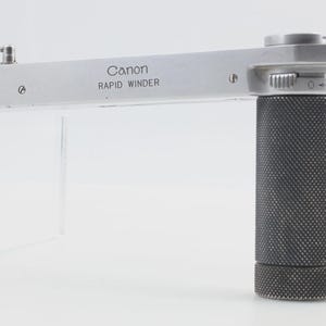 Puede incluir: Accesorio de cámara Canon Rapid Winder plateado. El dispositivo tiene un agarre negro texturizado y un brazo plateado con el logotipo de Canon. El Rapid Winder está diseñado para avanzar la película rápidamente. El accesorio está diseñado para acoplarse a un cuerpo de cámara.