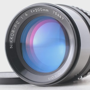 May include: A black Nikon Nikkor-PC 1:4 f=200mm camera lens with a lens cap. The lens has a blue and pink reflective surface. The lens has the text "NIKKOR-PC 1:4 f=200mm 75447" printed on it.