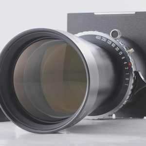 Op de afbeelding: Een grootformaat cameralens met een zwarte en zilveren afwerking. De lens heeft een groot diafragma en een reeks genummerde instellingen. De lens is gemonteerd op een zwarte rechthoekige basis.