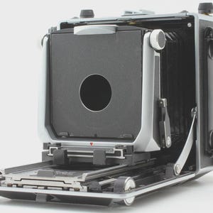 May include: A vintage large format camera with a black bellows and a silver-toned body. The camera has a large lens opening and a ground glass focusing screen. The camera is open, revealing its internal components.