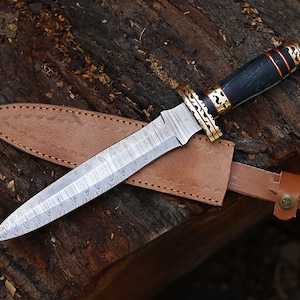May include: A Damascus steel dagger with a patterned blade and a black handle with gold accents. The dagger rests in a brown leather sheath with a belt loop, displayed on a wooden surface.