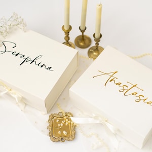 May include: Two ivory gift boxes, one with "Seraphina" in black script and the other with "Anastasia" in gold script. The boxes are tied with ivory ribbons and decorated with white flowers and pearls. Three lit candles in gold holders are in the background.