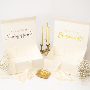 May include: Two open ivory gift boxes with the text "WILL YOU BE MY Maid of Honor?" and "Bridesmaid?" in gold and black script. The boxes are decorated with candles, flowers, and ribbons, perfect for a wedding.