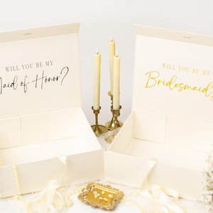 May include: Two open ivory gift boxes with the text "WILL YOU BE MY Maid of Honor?" and "WILL YOU BE MY Bridesmaid?" in elegant script. The boxes are decorated with flowers, candles, and a small decorative tray.