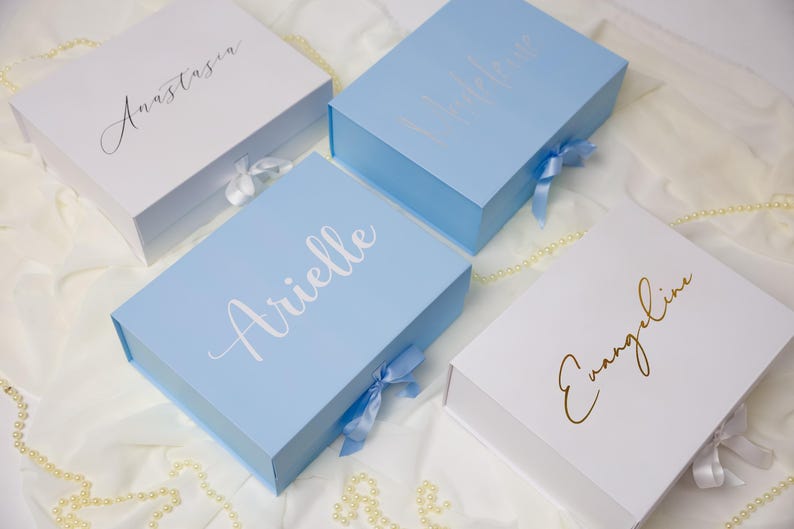 May include: Four rectangular gift boxes with magnetic closures and satin ribbon ties. Two boxes are white with black and gold script text reading Anastasia and Evangeline. Two boxes are light blue with white and silver script text reading Arielle.