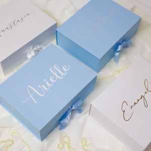 May include: Four gift boxes in white and light blue, each with a name in cursive script. The white boxes have gold lettering, while the blue boxes have white lettering. Each box has a matching ribbon.
