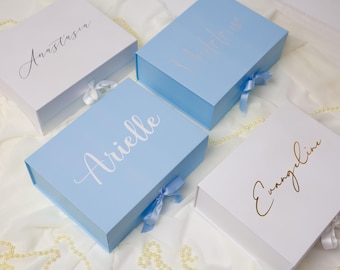 May include: Four rectangular gift boxes with magnetic closures and satin ribbon ties. Two boxes are white with black and gold script text reading Anastasia and Evangeline. Two boxes are light blue with white and silver script text reading Arielle.