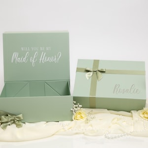 May include: Two sage green rectangular gift boxes. One box is open, displaying the text "WILL YOU BE MY Maid of Honor?" on the inside lid. The second box is closed, tied with a matching ribbon, and features the name "Rosalee" printed on the front.