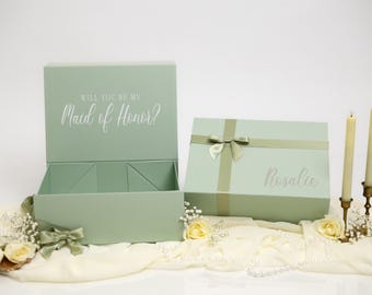 May include: Two sage green rectangular gift boxes. One box is open, displaying the text "WILL YOU BE MY Maid of Honor?" on the inside lid. The second box is closed, tied with a matching ribbon, and features the name "Rosalee" printed on the front.