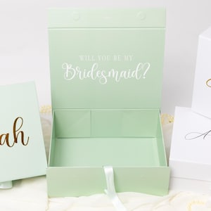 May include: A collection of rectangular gift boxes in light green and white. One open light green box features the white text Will You Be My Bridesmaid? while others display gold or black script names like Delilah, Evangeline, and Anastasia.