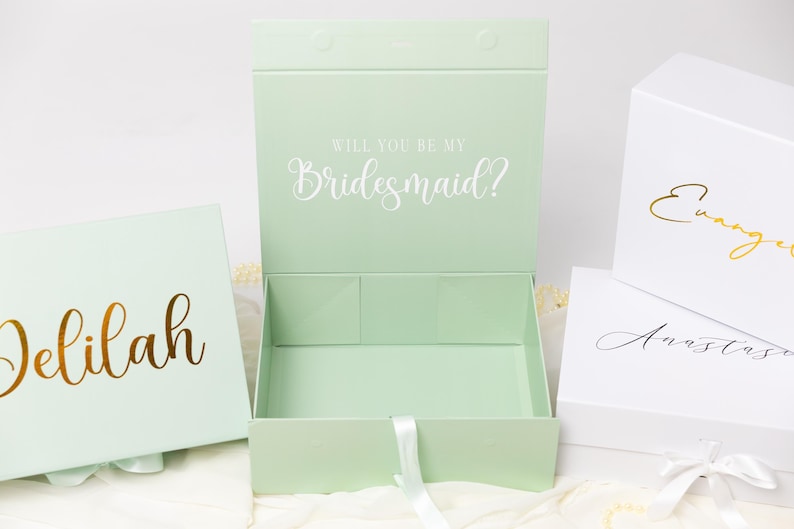 May include: A collection of rectangular gift boxes in light green and white. One open light green box features the white text Will You Be My Bridesmaid? while others display gold or black script names like Delilah, Evangeline, and Anastasia.