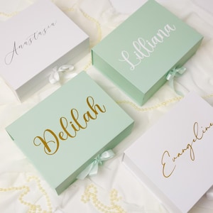 May include: Four rectangular gift boxes with magnetic closures and ribbon ties. Two boxes are white and two are light green. Each box features a custom name in cursive script: Anastasia, Lilliana, Delilah, and Evangeline, rendered in gold or white.