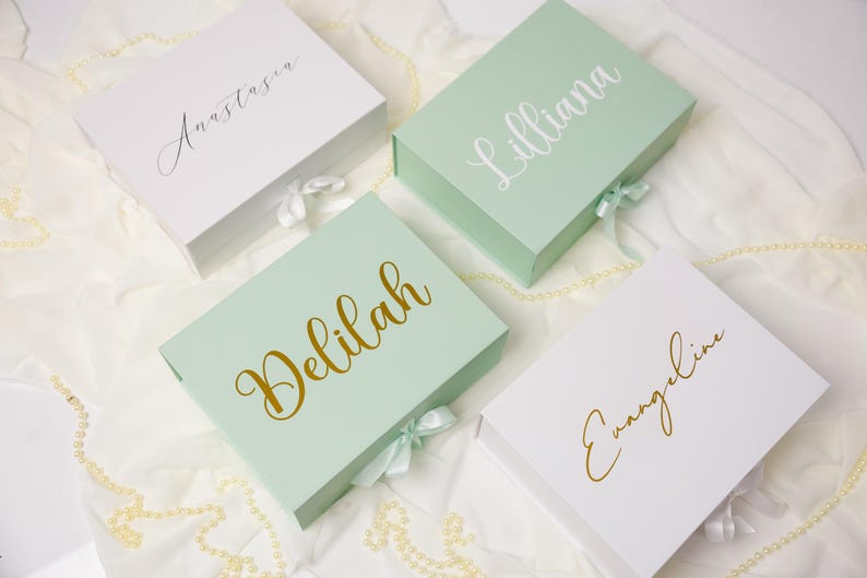 May include: Four rectangular gift boxes with magnetic closures and ribbon ties. Two boxes are white and two are light green. Each box features a custom name in cursive script: Anastasia, Lilliana, Delilah, and Evangeline, rendered in gold or white.