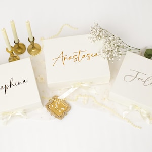 May include: Three square white gift boxes with ribbon closures, each personalised with a name in script lettering: Seraphina, Anastasia, and Juliette. The boxes are arranged with three thin cream candles in gold holders and sprigs of baby's breath.